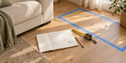 Measuring tape and floor-plan notebook beside an ivory sofa, with a furniture footprint marked on an oak floor